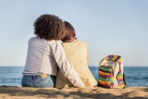 Image of a lesbian couple embracing on the beach next to a rainbow colored backpack. Showing the type of relationship you can have with online marriage counseling and online couples therapy in North Carolina. You can even get specialized LGBT couples counseling on in Chapel Hill, North Carolina.
