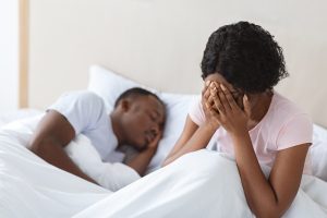 Image of a crying Black woman sitting her bed next to her husband. Representing a couple that could benefit from online marriage counseling in Chapel Hill, North Carolina. In situations like this online couples therapy in North Carolina can help reduce conflict.