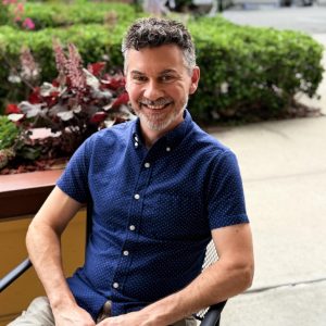 Licensed psychologist Dr Chris Weintrob seated outdoors on a patio, smiling, wearing a navy blue short-sleeve button-down shirt. Provides psychological evaluations for children, adolescents, and adults.